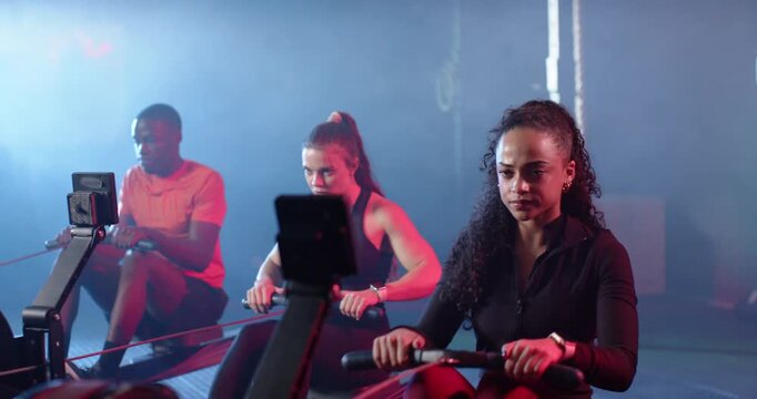 Diverse workout partners gripping rowing handles, rowing on machines in dim studio for endurance