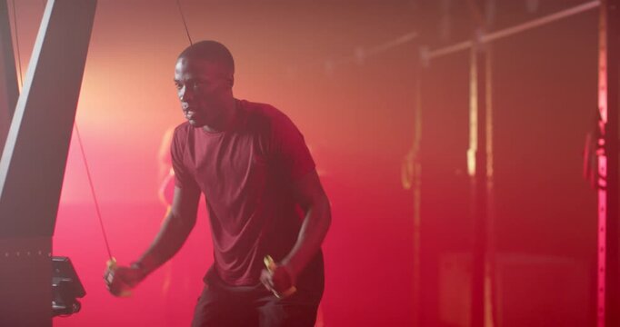 Adult African American man in gym in maroon tee gripping rope handles, swinging by frame, training