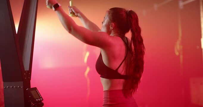 Female athlete gripping overhead handles and pulling cables for strength on gym rig under red haze
