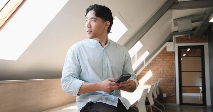 Asian man receiving smartphone notification scrolling on bench in loft glancing at skylight smiling