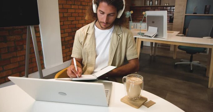 Headphones playing lecture, adult man writing in notebook using open laptop and mug in coworking