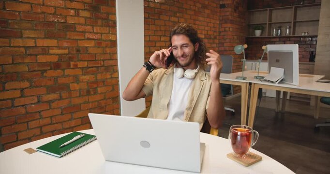 Man answering incoming phone call, adjusting headphones, checking laptop in brick office for work