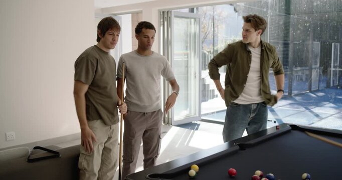 Diverse male friends at home examining racked balls, pointing cues and taking shot to play pool