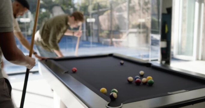 Diverse male friends in glass room playing pool, cue striking and scattering balls on black table