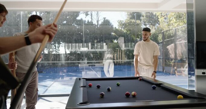 Diverse male friends striking cue, sending balls across black-felt pool table by glass wall for fun