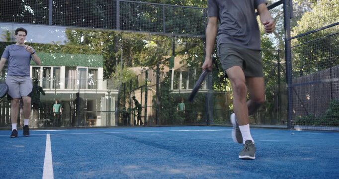 Diverse male friends playing on padel court paddles low shot causing lunging falling mate helping