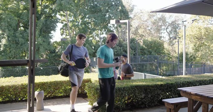 Four male friends leaving court, carrying paddles, gathering at picnic table, chatting