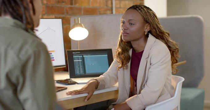 African American team colleague gesturing, seated woman at desk pointing monitor clarifying chart