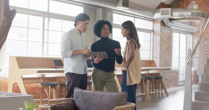 Diverse coworkers gathering, center turning tablet, left tapping screen and discussing work in loft