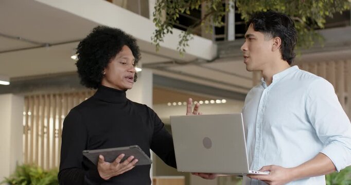 Diverse male coworkers gesturing and discussing for work, holding tablet and silver laptop, walking