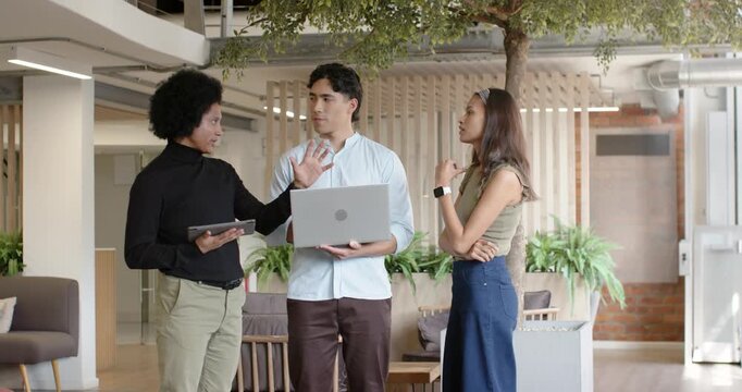 Diverse team planning in lobby left raising hand tablet center reading laptop right gesturing watch