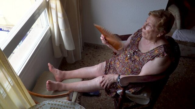 An elderly woman fanning herself on a hot afternoon, sitting in a chair at home 