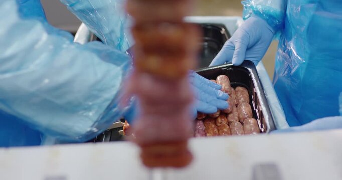 Gloved hands arranging small raw sausages on wooden skewers in black plastic trays at a food production facility
