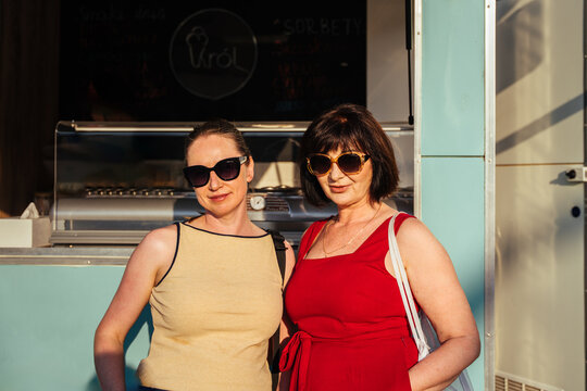Stylish women friends in sunglasses posing by ice cream kiosk on sunny summer day