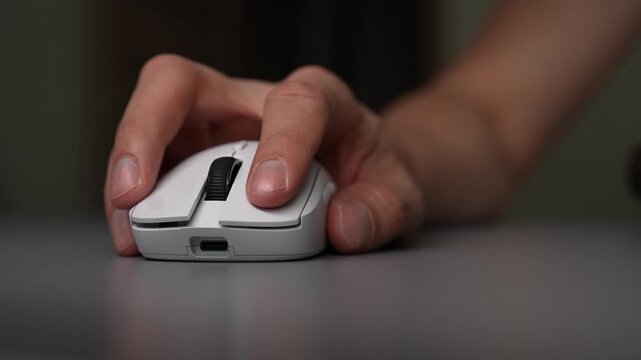 Close-up cropped shot of person hand using modern white wireless mouse on grey desk. Fingers move between buttons and scroll wheel, clicking and navigating. Technology and office work concept.