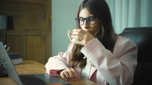 Young Hispanic woman working in her office while drinking a cup of coffee, focusing on her tasks at a desk and enjoying a brief moment of refreshment during her workday.
