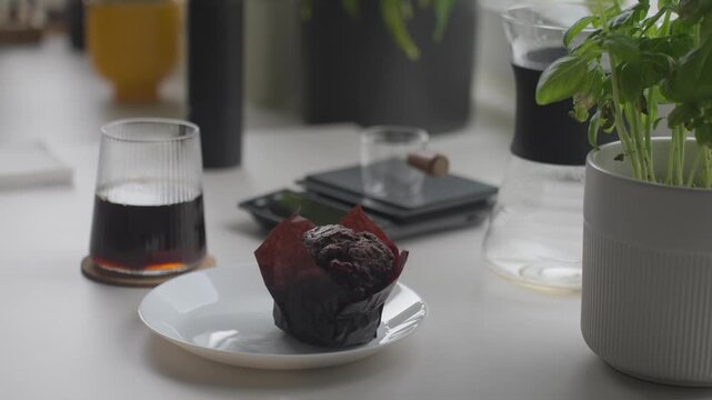 Hand places chocolate muffin on white plate beside glass of coffee on kitchen counter. Concept of coffee break, dessert and simple snack