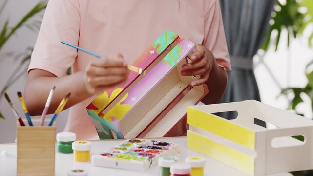 Creative female artist applying vibrant acrylic paints onto a handmade wooden crate. Concept of DIY home decor, artistic hobby, art therapy, and domestic leisure.