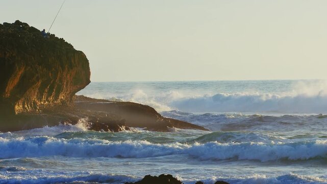 Cinematic 4K slow motion footage of powerful ocean waves crashing against a rugged rocky cliff where a lone fisherman stands. The scene captures the raw beauty of the coastal landscape bathed in the s