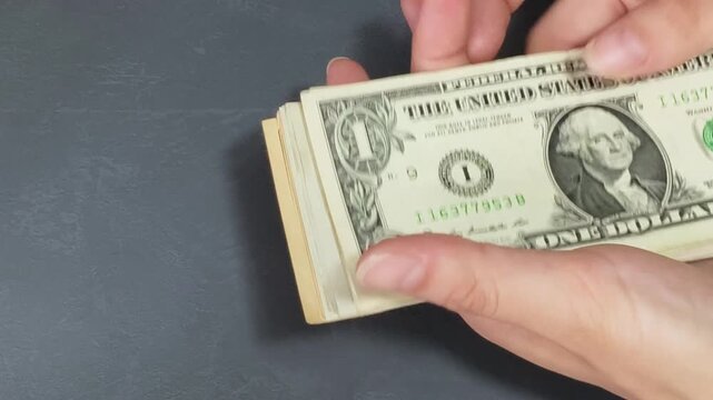 Close-up of hands gathering and taking away a scattered stack of small denomination US dollar bills from a graphite grey table.
