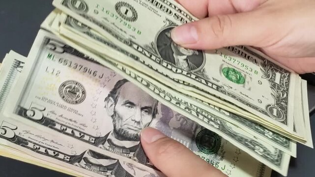 Close-up of hands counting small denomination US dollar bills with a calculator partially visible on a graphite grey background.