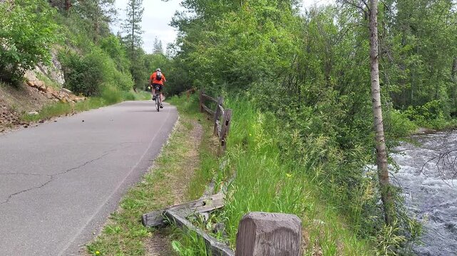 Bicycle rider in orange riding away from camera down paved trail near downed split-rail fence above cree