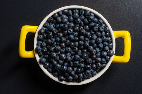 Fresh blueberries in white bowl with yellow handles, closeup. Top view of round ceramic pot filled with ripe fresh blueberries on dark background. Healthy organic summer berries food photography