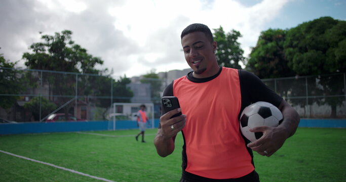 Smiling Latin Hispanic man of African descent checking smartphone while holding soccer ball on green field during friendly match break, digital connection, modern sport lifestyle