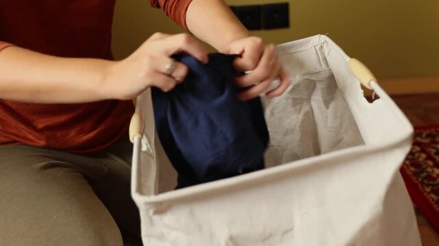 Close up of woman putting dark blue clothes into textile laundry basket. Female hands sorting dirty clothing at home, domestic routine concept