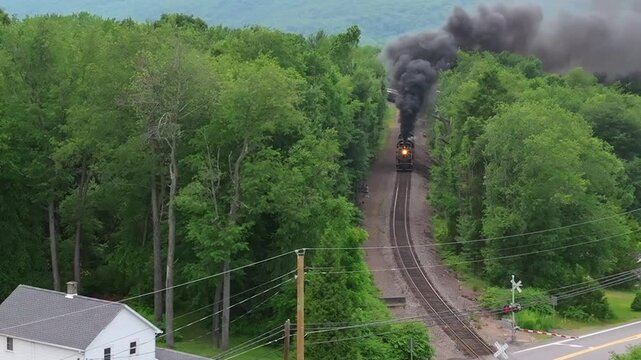 A Steam train moves along its tracks through a lush forest area close to a small town. Smoke rises from the train as it journeys through the landscape.