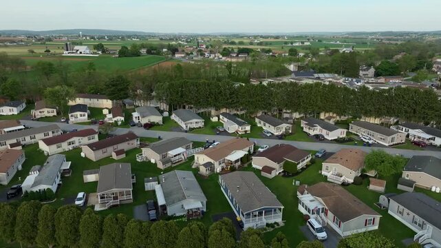 The camera shows a neighborhood with mobile, manufactured homes and roads during early evening. In the background, there are green fields and hills. The view captures the layout of the area well.