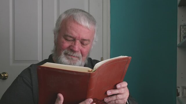 An older man sits in a room reading a book. He turns the pages slowly and shows different emotions as he engages with the text. The light comes through the window, making it a nice time to read.