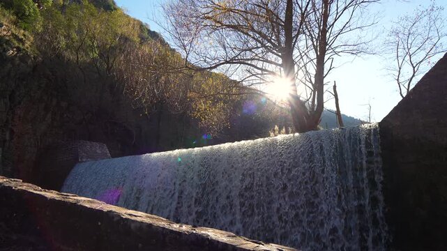 Low angle static shot of a stone waterfall dam with sun rays breaking through tree branches