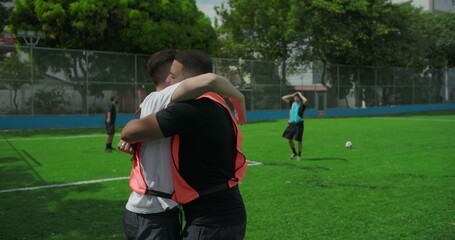 Obraz premium Soccer teammates embrace and raise fists in celebration after scoring goal on soccer field, shouting with joy, while kneeling opponent watches, showing unity