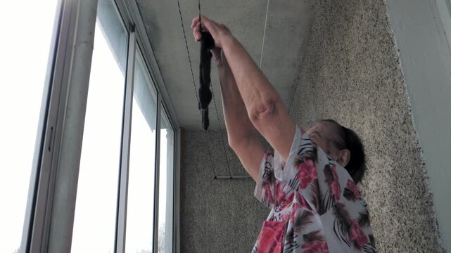 Senior woman hanging wet socks on a clothesline on her apartment balcony. Showcasing simple and traditional domestic chore performed by an older person