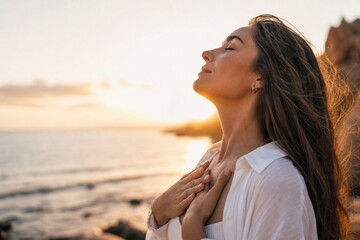 Obraz premium Beautiful young woman with closed eyes holding hands on her chest while practicing mindfulness and deep breathing on the beach during a golden sunset