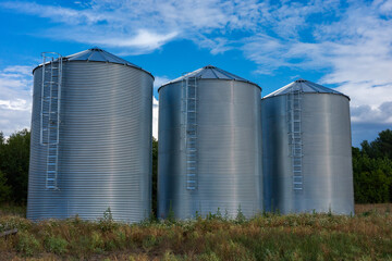 Three modern corrugated steel grain silos standing in a row on a farm field. Agricultural storage tanks with ladders under a blue sky with white clouds. Industry and farming concept. © Fotoforce