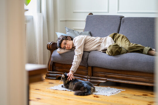 Tranquil careless woman lazy lying on couch enjoying peaceful living in company of domestic kitty, passive calm female petting cat while resting on sofa, no stress and rush, mindful procrastination.