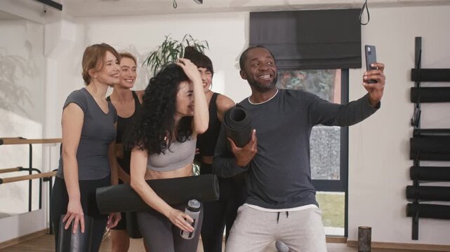 Group of yoga people having fun during break. Multicultural people smiling at camera at yoga studio. African American man holding phone and taking selfie of yoga group. Peope holding fitness mats.