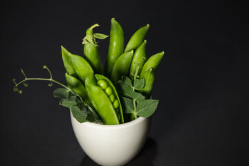 Fresh raw green pea pods with leaves in a white cup on a dark background © Denis