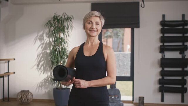 Cheerful woman standing at yoga studio. Smiling female wearing sportclothing and holding mat. Woman preparing for yoga session. Female practicing meditation class. Yoga instructor smiling at camera.