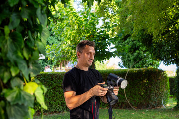Fototapeta premium Behind the scenes, a photographer in a black shirt adjusts a professional camera outdoors in a green garden, focusing intently before capturing the shot