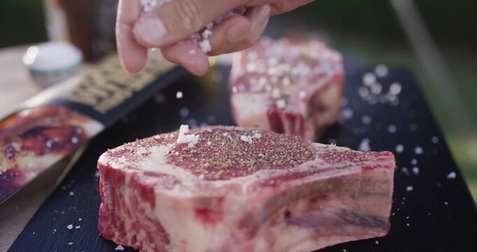 Coarse salt flakes fall onto a raw seasoned steak on a dark board before grilling outdoors