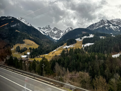 A dramatic alpine landscape highlights towering, jagged mountain peaks covered in patches of snow under a heavy, overcast winter sky. Dense green pine and fir forests blanket the steep valley walls, s