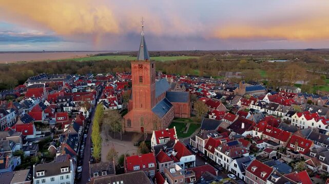 Aerial wide angle panoramic view of a majestic historic brick church cathedral with a tall spire tower standing in the center of a medieval old town under a dramatic sunset sky
