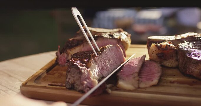 Knife slicing a grilled steak held by a carving fork on a wooden cutting board in warm evening light