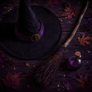 Witch hat and broom with purple potion bottle and autumn leaves on dark wood table.