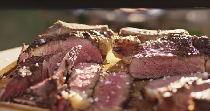 Salt flakes falling onto sliced grilled steak resting on a wooden cutting board in warm sunlight