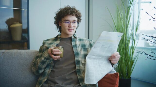 Man sitting on a couch, reading a newspaper and drinking from a glass in a modern indoor space during daytime
