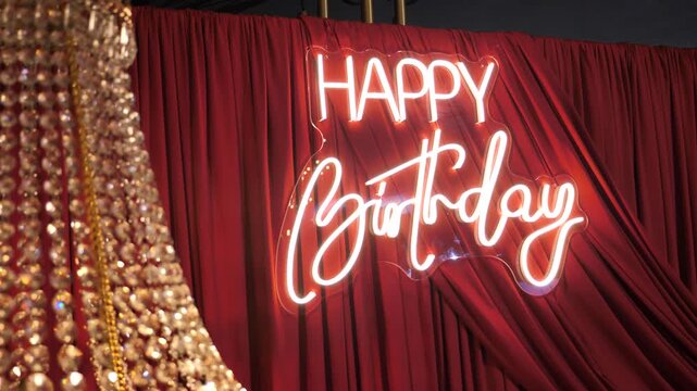 A glowing white neon sign with Happy Birthday inscription mounted on a luxury dark red fabric drapery with a golden crystal chandelier blur in the foreground.